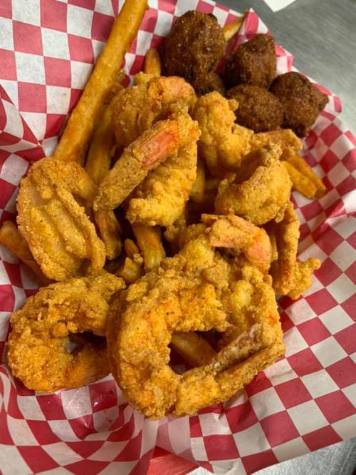 A basket of fried shrimp, French fries, and two hushpuppies on a red and white checkered paper.