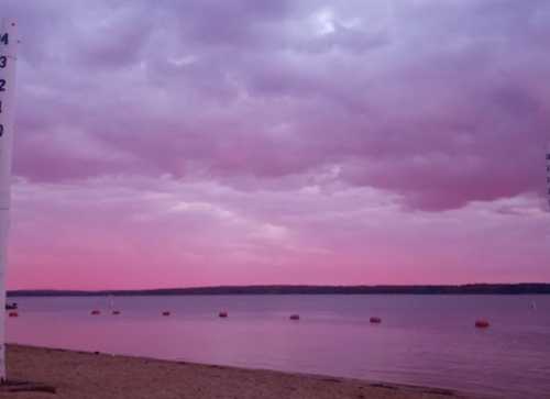 A serene beach scene at sunset, with pink clouds reflecting on calm water and buoys floating in the distance.