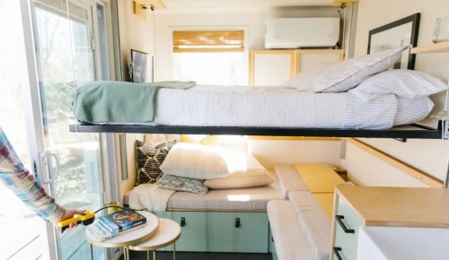 A compact, modern living space with a foldable bed, cozy seating, and natural light from large windows.