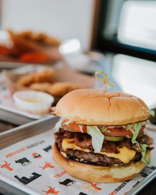 A juicy cheeseburger with lettuce, tomato, and bacon, served on a tray with dipping sauce in the background.