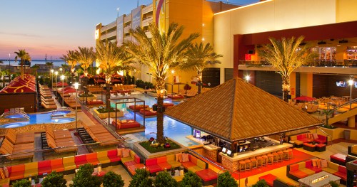 A vibrant resort pool area with palm trees, lounge chairs, and a tiki bar, set against a sunset sky.