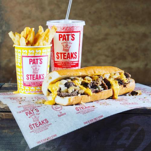 A cheesesteak sandwich with melted cheese, fries in a cup, and a drink, all from Pat's King of Steaks.