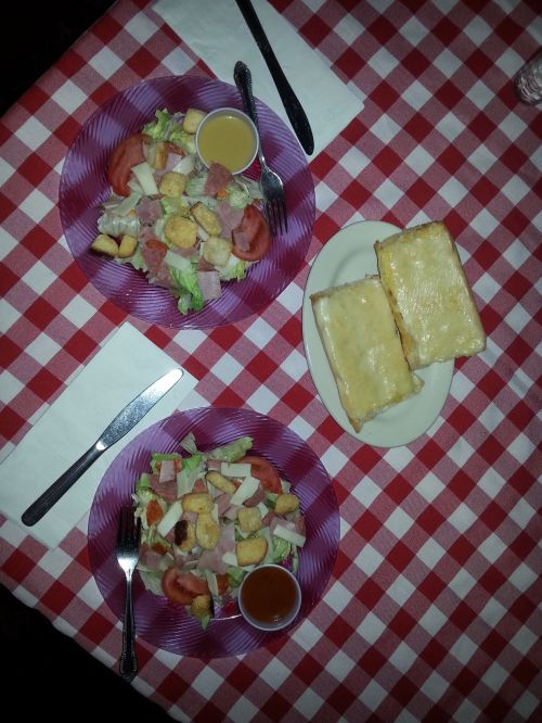 Two salads with croutons and dressing on a checkered tablecloth, accompanied by two slices of melted cheese bread.