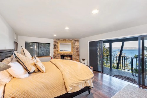Cozy bedroom with a large bed, stone fireplace, and balcony overlooking a scenic lake view.
