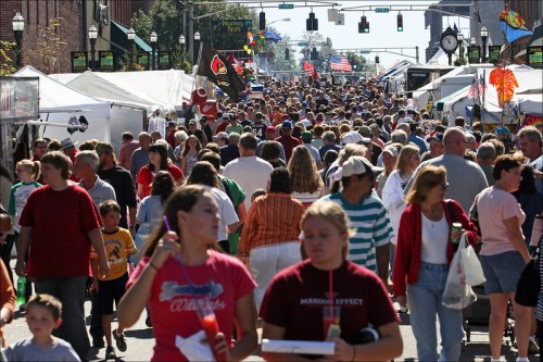 A bustling street fair with large crowds, tents, flags, and people enjoying the festivities on a sunny day.