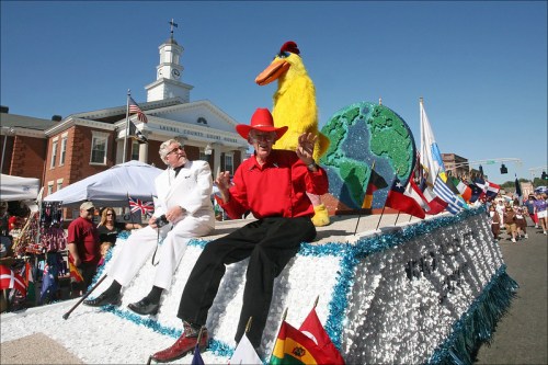 A colorful parade float featuring a man in a red shirt and cowboy hat, alongside a large yellow bird and flags from various countries.