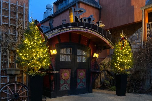 A festive entrance with two lit Christmas trees flanking a colorful door, set against a twilight sky.