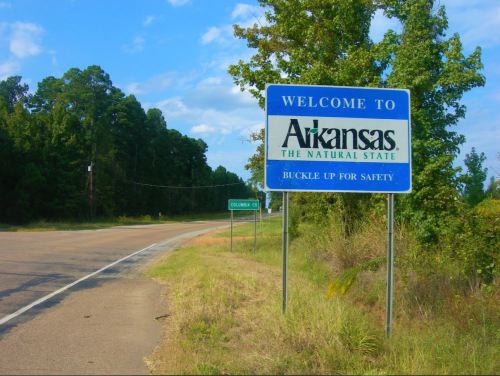 Welcome sign for Arkansas, "The Natural State," with a reminder to buckle up for safety, alongside a rural road.