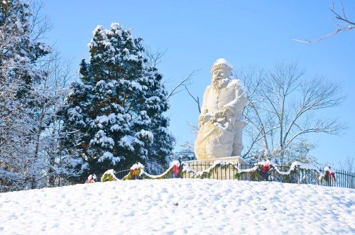 A snow-covered statue of a figure holding a child, surrounded by trees and festive decorations in a winter landscape.