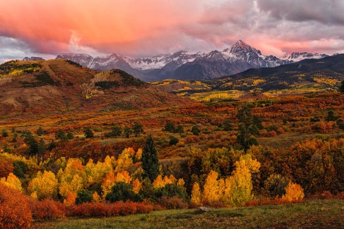 A vibrant autumn landscape with colorful trees, mountains, and a dramatic sunset sky.