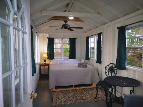 Cozy interior of a small cabin with a bed, table, and large windows, featuring white walls and dark curtains.