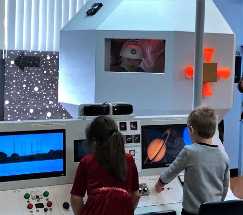 Children interact with a space-themed exhibit featuring screens and a model spacecraft in a bright, educational setting.