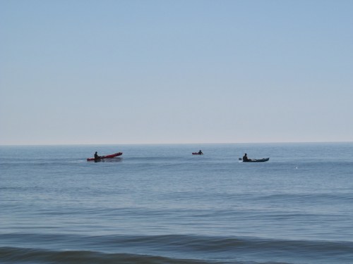 Three kayakers paddle on a calm ocean under a clear blue sky.