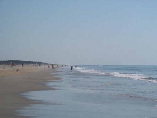 A serene beach scene with people walking along the shore and gentle waves lapping at the sand under a clear sky.