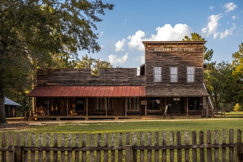 Historic wooden drug store with a red roof, surrounded by greenery and a wooden fence under a blue sky.