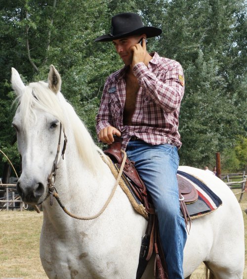 A man in a plaid shirt and cowboy hat talks on a phone while riding a white horse in a rural setting.