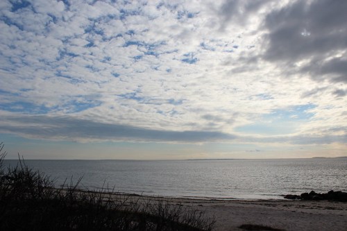 A serene beach scene with a cloudy sky and calm water, framed by distant land and gentle waves.