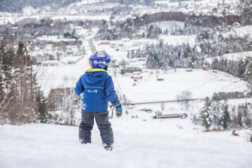 A child in winter gear stands on a snowy hill, overlooking a snow-covered town and landscape below.