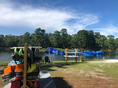 A serene lake scene with colorful life jackets on display, inflatable water toys in the background, and trees surrounding the area.