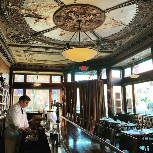 A vintage bar interior with ornate ceiling details, a bartender preparing drinks, and large windows letting in natural light.
