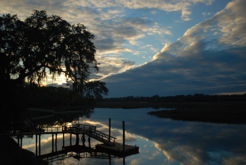 A serene sunset over a calm river, with a wooden dock and trees silhouetted against a colorful sky.