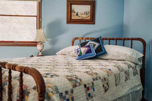 A cozy bedroom with a wooden bed, quilted blanket, decorative pillows, and a lamp beside a window.