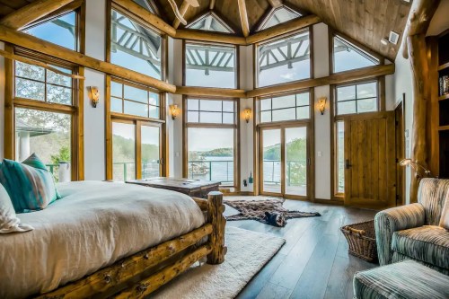 Cozy bedroom with large windows, wooden beams, and a view of the lake, featuring a bed and a comfortable chair.