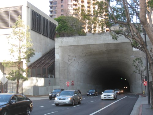 A concrete tunnel entrance with cars approaching, surrounded by trees and buildings in an urban setting.