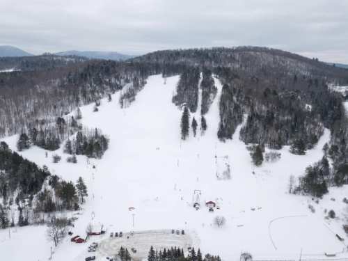 Aerial view of a snowy ski slope surrounded by trees and mountains under a cloudy sky.