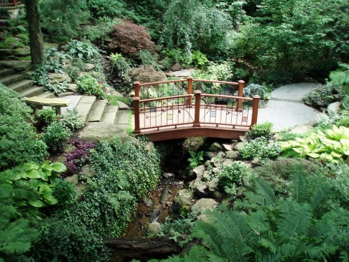 A serene garden with lush greenery, a wooden bridge over a small stream, and winding stone pathways.