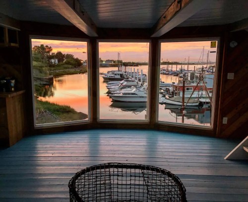 A cozy room with large windows overlooking a marina at sunset, featuring boats and calm water reflections.