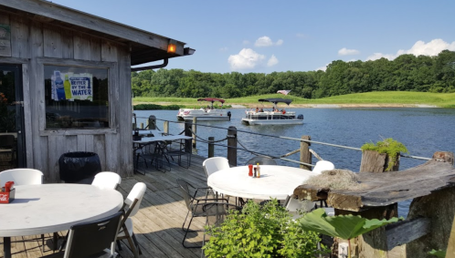 A wooden deck by the water with tables, overlooking boats on a calm lake and green hills in the background.