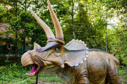 A lifelike dinosaur sculpture of a triceratops in a lush, green outdoor setting.