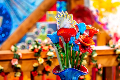 A vibrant arrangement of colorful glass flowers in a blue vase, set against a bright, festive background.