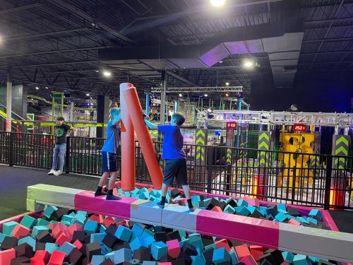 Two kids play on a foam pit, balancing on a beam while holding large foam poles in a colorful indoor trampoline park.