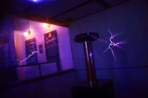 A Tesla coil emits purple lightning bolts in a dimly lit room with warning signs on the walls.