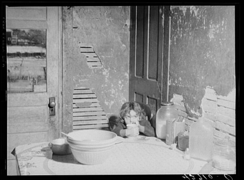 A child sits at a table in a rustic room, holding a cup, with bowls and jars nearby. The walls show signs of wear.