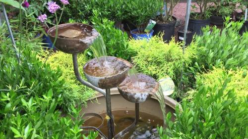 A tiered water fountain with three bowls, surrounded by lush green plants in pots.