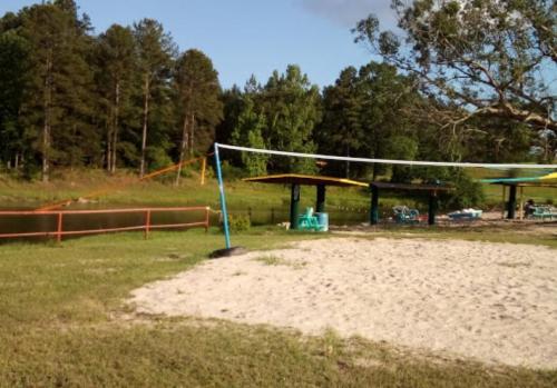 A sandy volleyball court near a pond, surrounded by trees and picnic areas with colorful canopies.