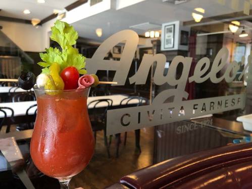 A vibrant Bloody Mary garnished with celery, olives, cherry tomatoes, and meats, set against a restaurant window.