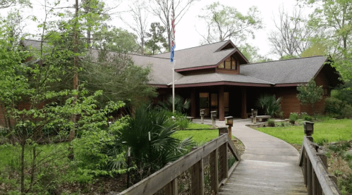 A wooden building surrounded by greenery, with a pathway leading to the entrance and an American flag in front.