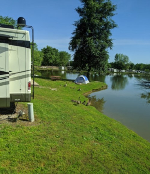 A camper parked by a serene lake, surrounded by green grass, trees, and a small tent nearby. Ducks are seen near the water.