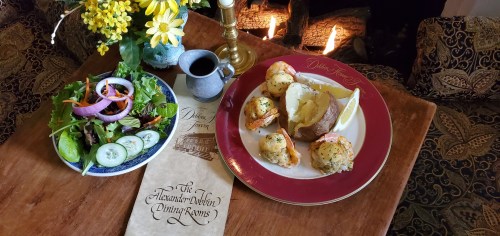 A plate with stuffed potatoes and shrimp, a fresh salad, and a menu on a wooden table near a cozy fireplace.