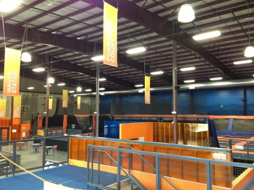 Interior of a trampoline park featuring various jumping areas, foam pits, and colorful banners hanging from the ceiling.