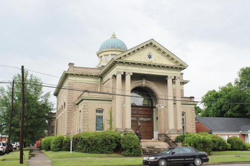 Historic building with a dome, columns, and a star emblem, surrounded by greenery and a cloudy sky.