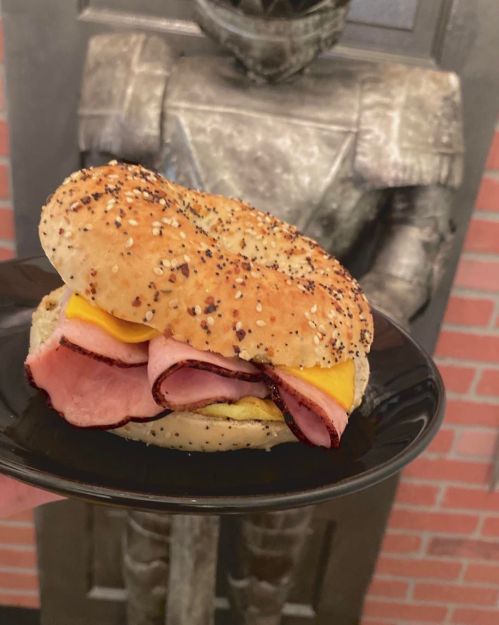 A sesame seed bagel filled with ham, cheese, and mustard, served on a black plate with a knight statue in the background.