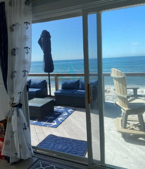 View of a beachside patio with outdoor furniture, ocean waves, and a clear blue sky through a sliding glass door.