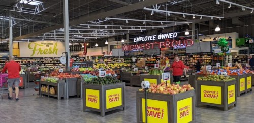 A spacious grocery store with colorful produce displays and signs promoting employee ownership and savings.