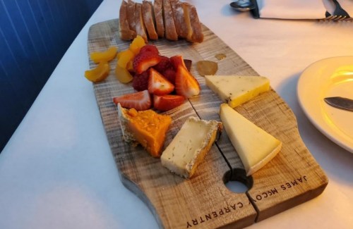A wooden cheese board featuring various cheeses, fresh strawberries, peaches, and slices of bread.