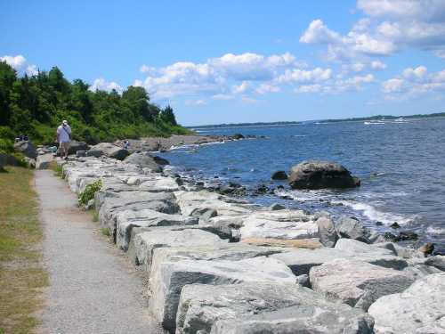 A rocky shoreline with a path, trees in the background, and a clear blue sky with scattered clouds.